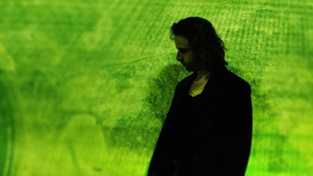 A dark-haired man in a black coat stands against a green-lit textured wall, gazing downward.