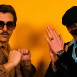 Funky District duo pose in retro-themed outfits with Nic Hanson wearing sunglasses against a yellow backdrop.