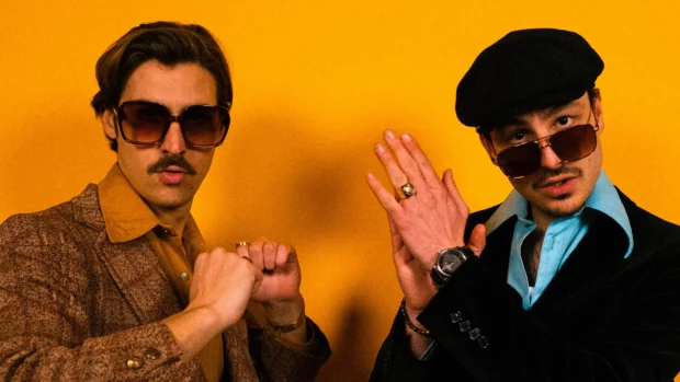 Funky District duo pose in retro-themed outfits with Nic Hanson wearing sunglasses against a yellow backdrop.