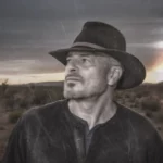 Man in a hat standing in a desert landscape at sunset, gazing into the distance.