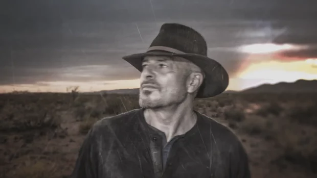 Man in a hat standing in a desert landscape at sunset, gazing into the distance.