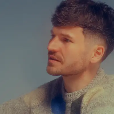 James Gruntz captured in a calm and thoughtful moment, seated casually on the floor in a pastel-patterned sweater and sneakers, against a soft gradient blue backdrop.