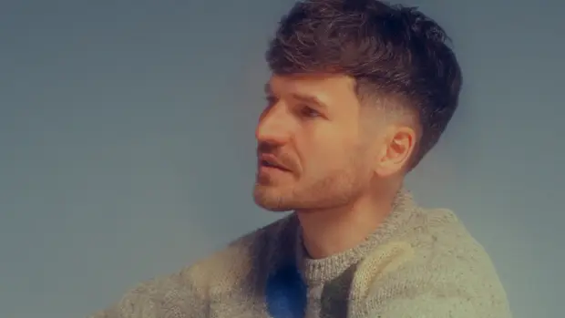 James Gruntz captured in a calm and thoughtful moment, seated casually on the floor in a pastel-patterned sweater and sneakers, against a soft gradient blue backdrop.