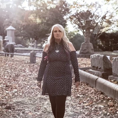 Jody Lynn walking through a cemetery with sunlight filtering through trees, evoking themes of resilience and reflection.