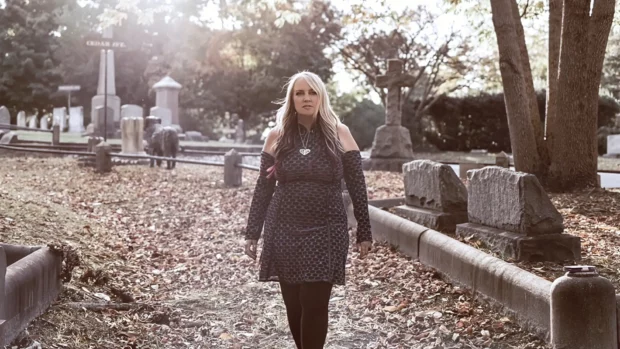 Jody Lynn walking through a cemetery with sunlight filtering through trees, evoking themes of resilience and reflection.