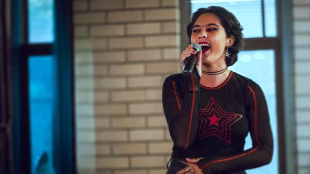 Jupiter Cubero performs passionately with a microphone, wearing a black star-themed outfit, set against a window-lit brick backdrop.