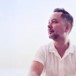 Milo A, seated against a soft, sunlit background, wearing a white shirt.