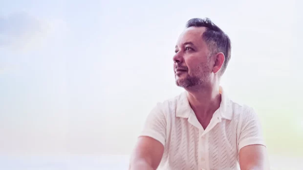 Milo A, seated against a soft, sunlit background, wearing a white shirt.