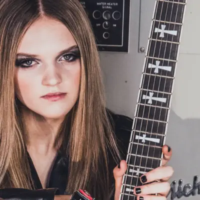 Sierra Levesque holding a black electric guitar, wearing black leather, sitting in front of a control panel.