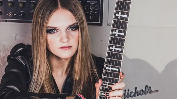 Sierra Levesque holding a black electric guitar, wearing black leather, sitting in front of a control panel.