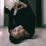 YME lying on the floor, making a heart gesture with her hands, wearing black attire, with a reflective mirror in the background.