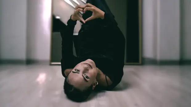 YME lying on the floor, making a heart gesture with her hands, wearing black attire, with a reflective mirror in the background.