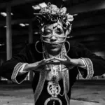 Black-and-white portrait of Yugen Blakrok wearing intricate jewelry and headpiece in a dimly lit industrial setting.