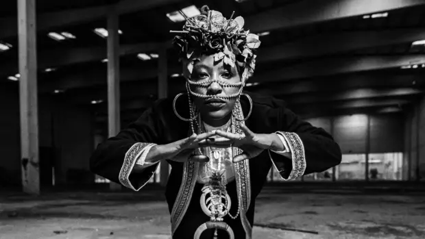 Black-and-white portrait of Yugen Blakrok wearing intricate jewelry and headpiece in a dimly lit industrial setting.